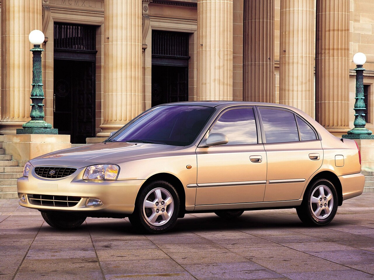 Hyundai Accent (II поколение, 1999 - 2012 г.в.): Золотистый седан на площади у здания с колоннами, вид спереди сбоку