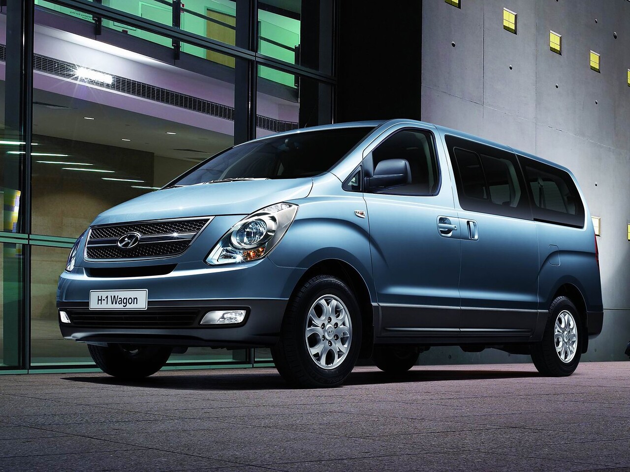 Hyundai H-1 (II поколение, 2007 - 2015 г.в.): Голубой минивэн у современного здания ночью, вид спереди сбоку