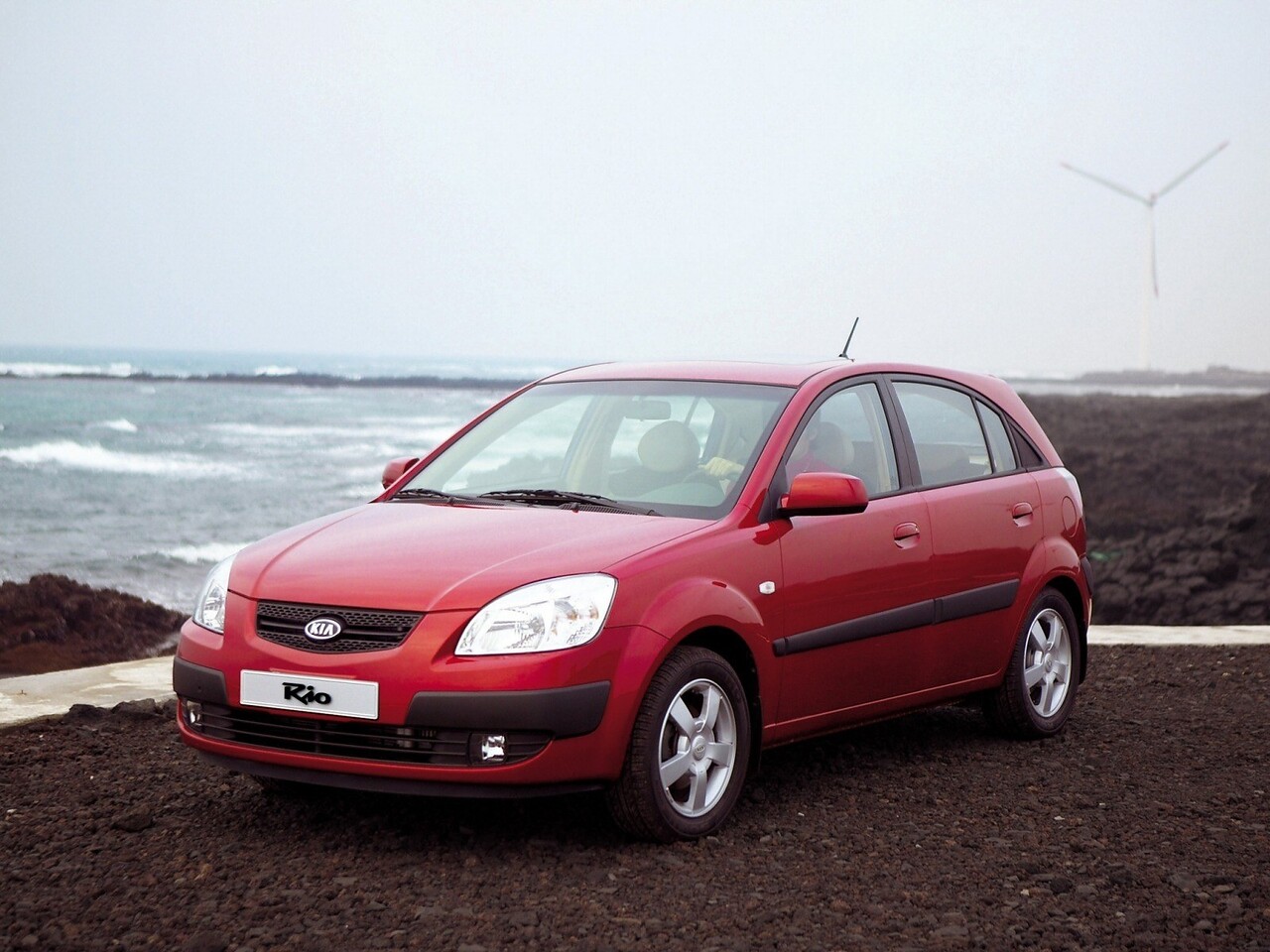 KIA Rio Хэтчбек (II поколение, рестайлинг, 2009 - 2011 г.в.): Три четверти спереди у побережья на фоне ветряного генератора