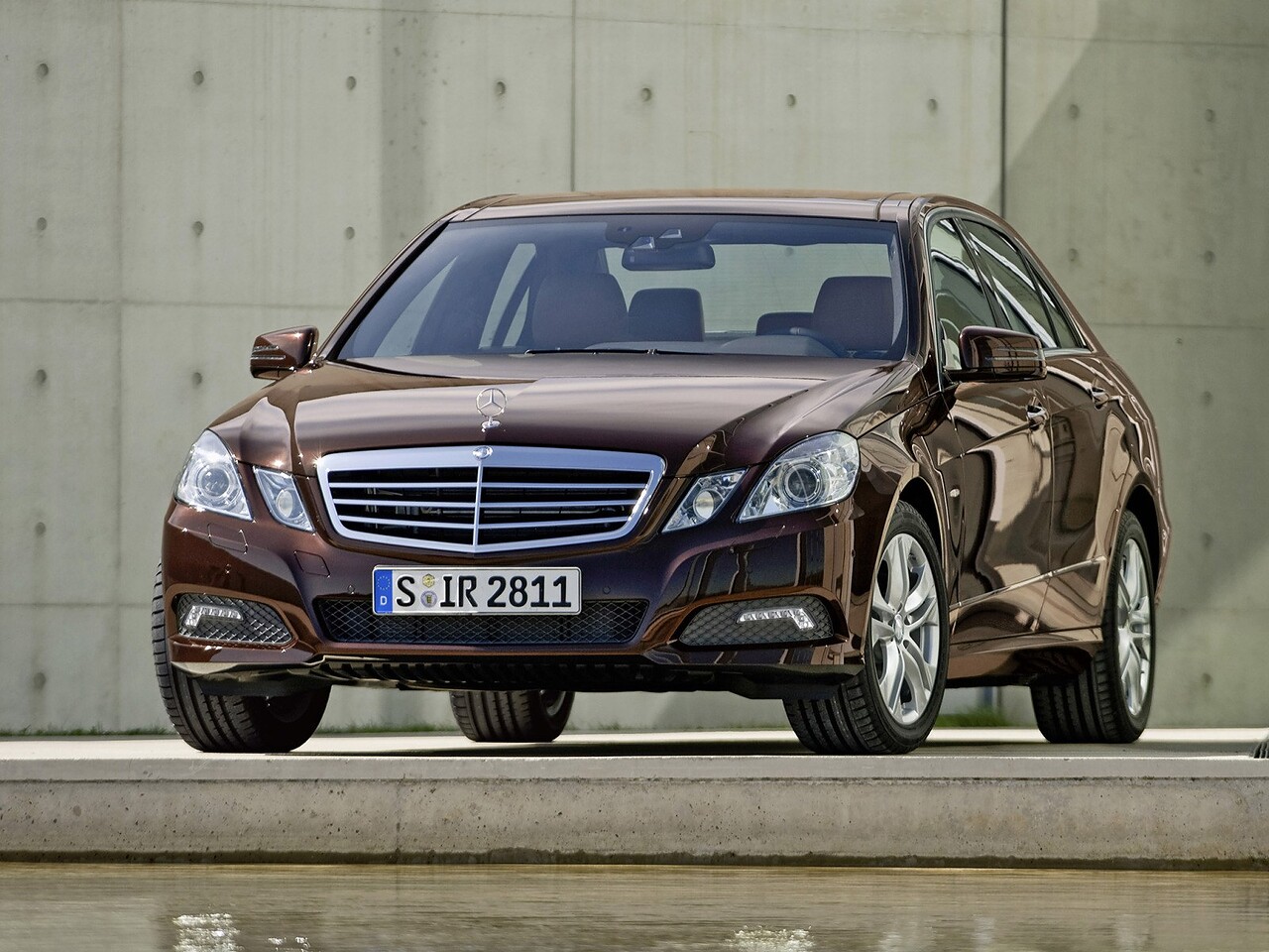 Mercedes-Benz E-Класс седан (w212, 2009 - 2012 г.в.): Коричневый седан у бетонной стены, вид спереди сбоку