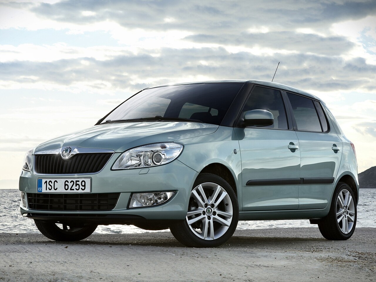 Skoda Fabia хэтчбек 5-дв. (II поколение, рестайлинг, 2010 - 2015 г.в.): Вид спереди в три четверти на грунтовой дороге под облачным небом