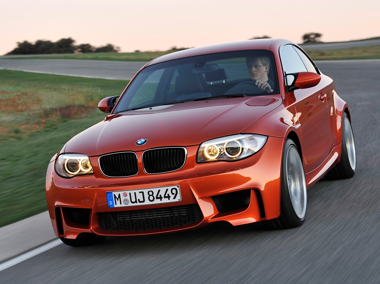 BMW 1-я серия М (E82, 2010 - 2014 г.в.): Оранжевое купе на извилистой дороге, вид спереди сбоку