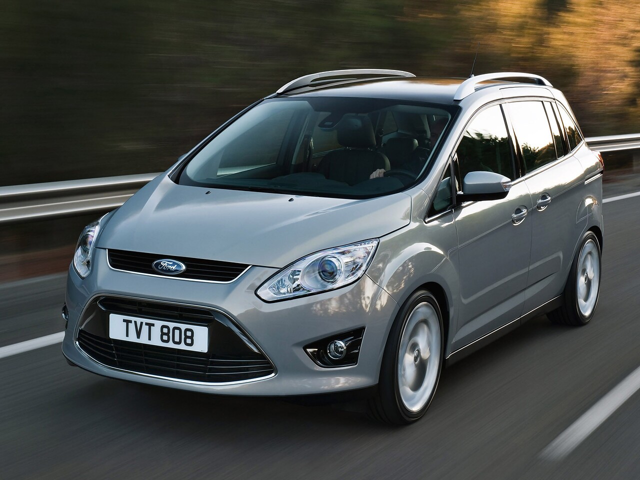 Ford Grand C-MAX 1.6 EcoBoost MT Titanium: Серебристый минивэн на дороге среди деревьев, вид спереди сбоку